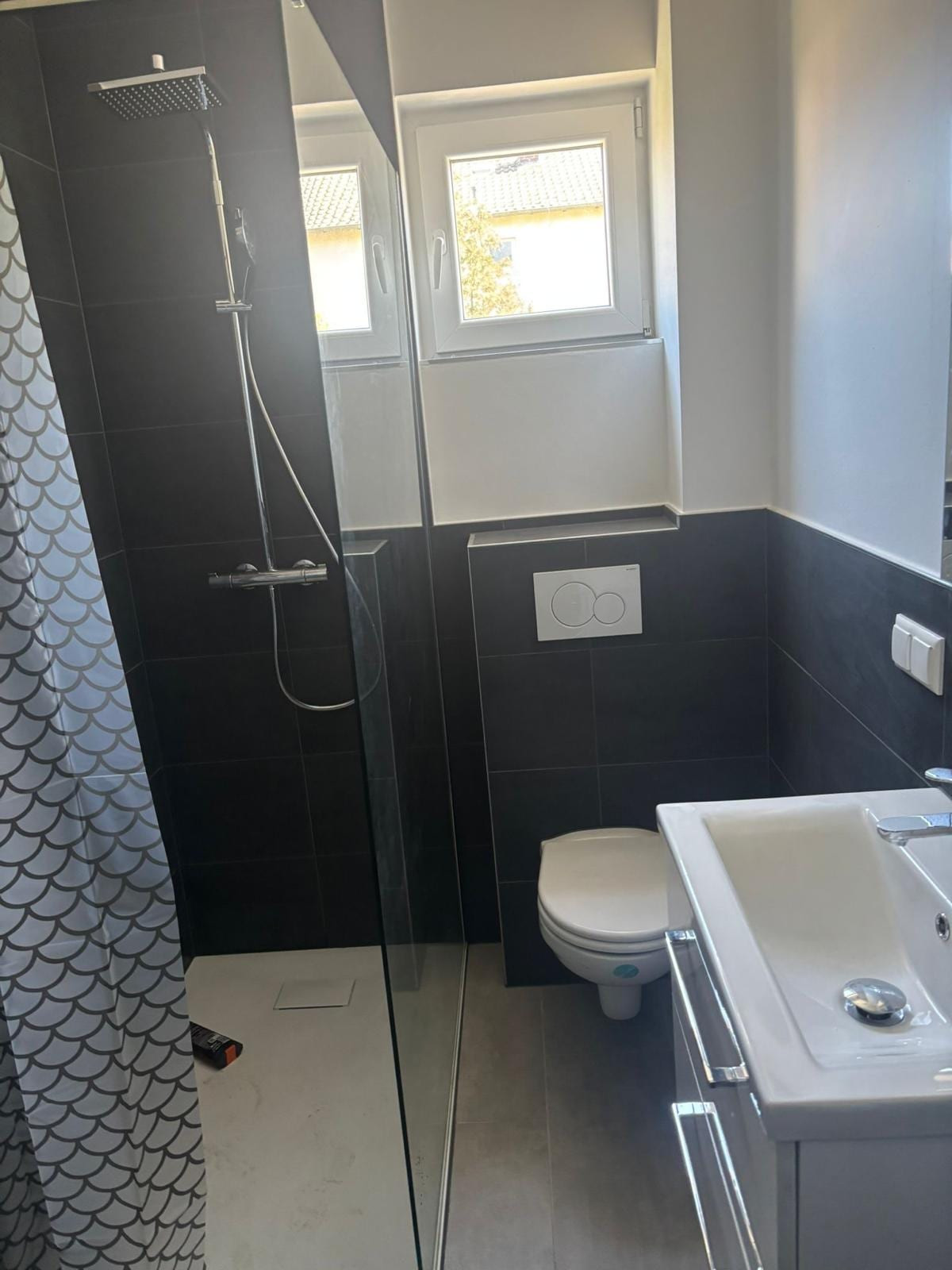 Kompaktes Badezimmer mit anthrazitfarbenen Fliesen, bodengleicher Glasdusche mit Regenbrause, wandhängendem WC und Waschtisch.