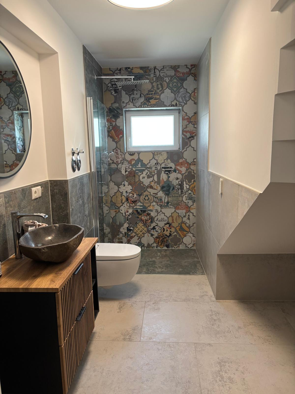 Gäste-Bad mit Naturstein-Aufsatzbecken auf Holz-Unterschrank, rundem Spiegel und einer bunten Collage-Fliesenwand in der Dusche.