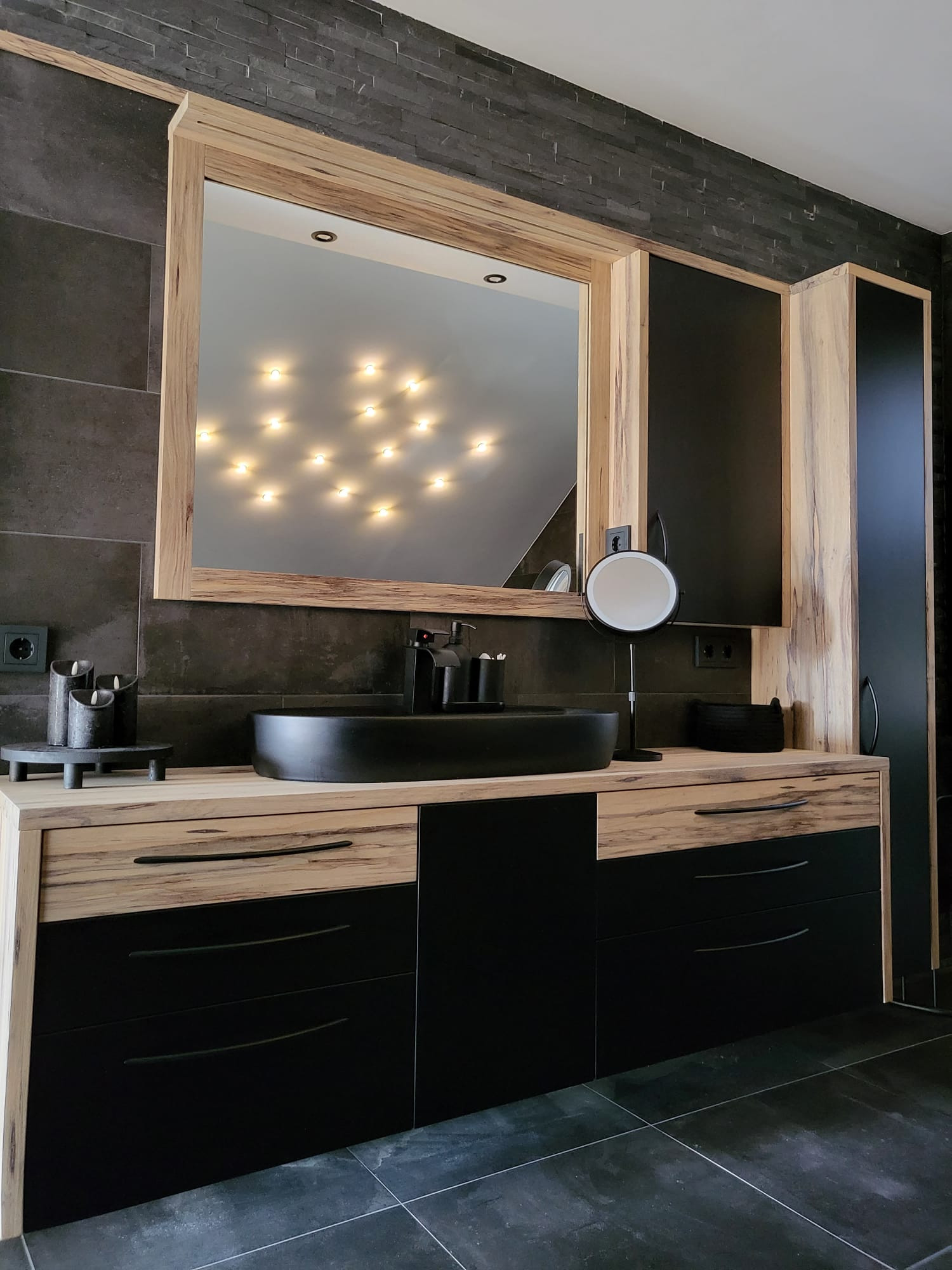 Modernes Badezimmer mit Holz-Waschtisch, dunkler Natursteinwand, mattschwarzem Aufsatzbecken und hinterleuchtetem Spiegel.