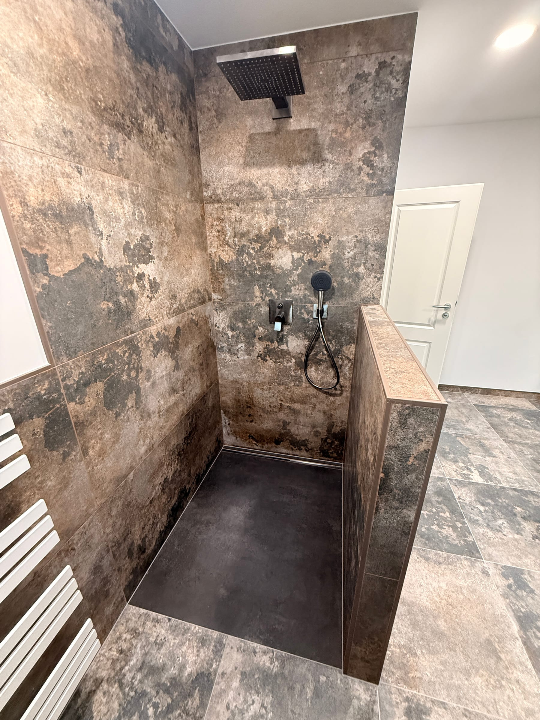Ebenerdige Dusche mit Regenbrause, schwarzer Armatur und Fliesen in Rostoptik, abgetrennt durch eine geflieste Halbwand.