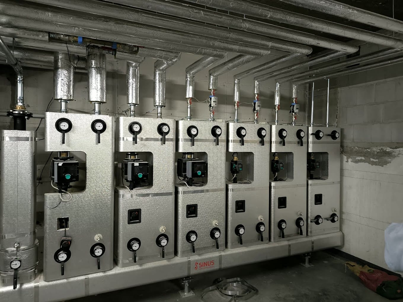 Technikraum mit mehreren silbernen Heizungs-Verteilerstationen, Manometern, Pumpen und gedämmten Rohrleitungen.